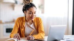 留着脏辫的年轻黑人妇女坐在书桌前，打开笔记本电脑，在纸和笔上写字 
